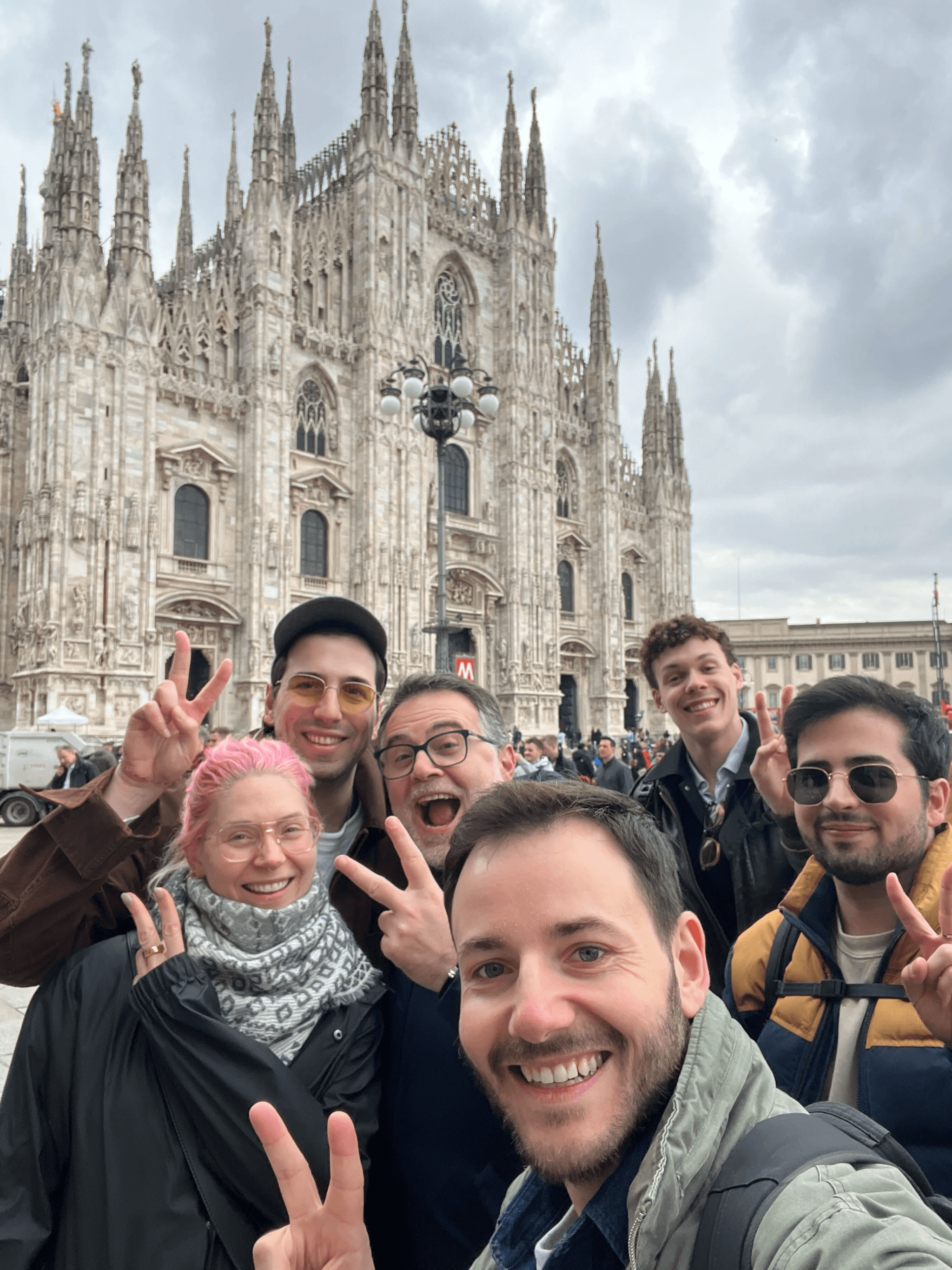 Six smiling teammates flashing peace signs in front of Milan Duomo